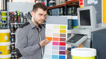 Junger Mann, der Farbe für das Malen unter Verwendung des Palettenschemas im Haushaltsgeschäft wählt Farbe Wunschfarbe Farbmischmaschine